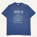 A blue short-sleeve graphic t-shirt featuring the text 'HÜSKER DÜ' and 'METAL CIRCUS' with a distressed circular emblem on the front, laid flat against a light off-white background.