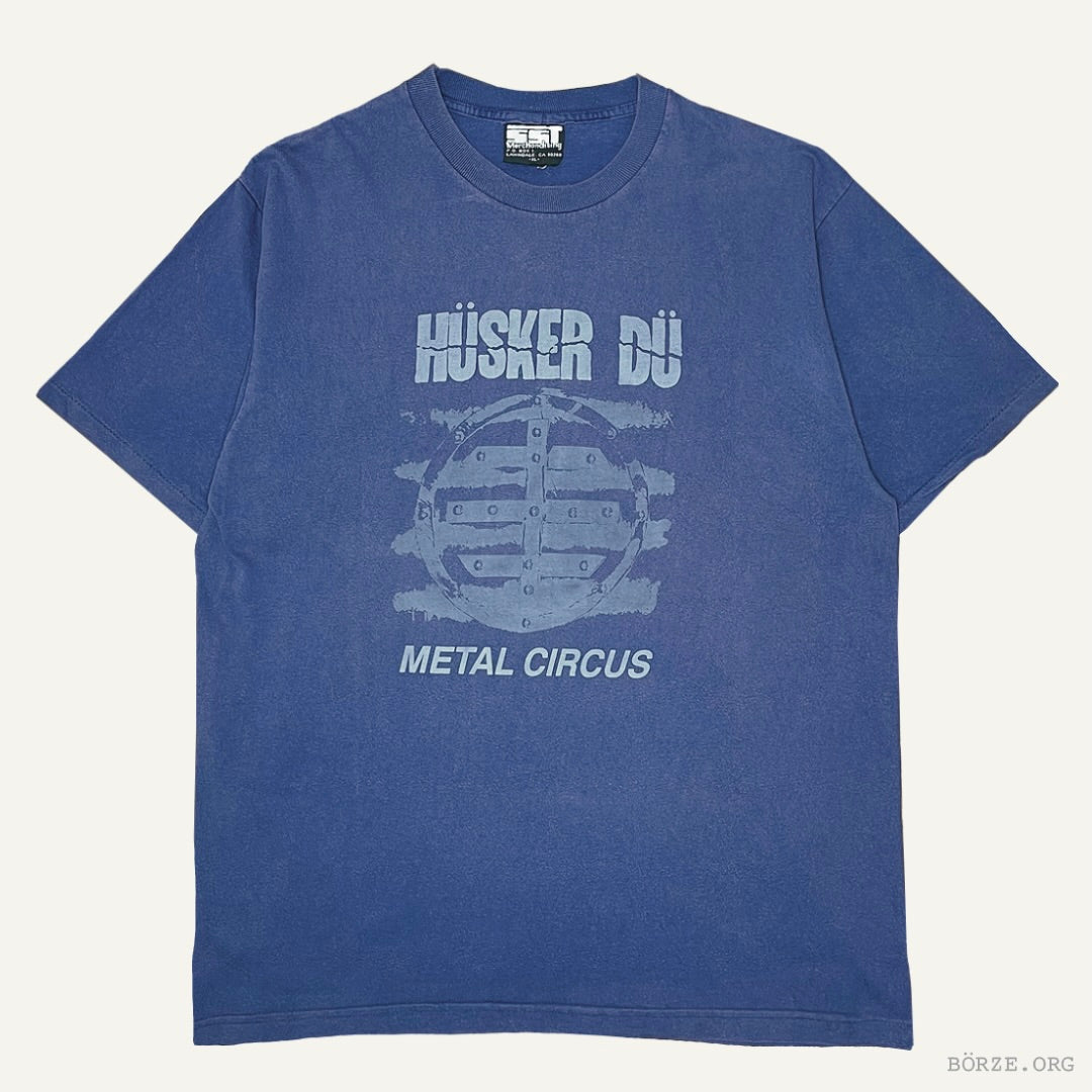 A blue short-sleeve graphic t-shirt featuring the text 'HÜSKER DÜ' and 'METAL CIRCUS' with a distressed circular emblem on the front, laid flat against a light off-white background.